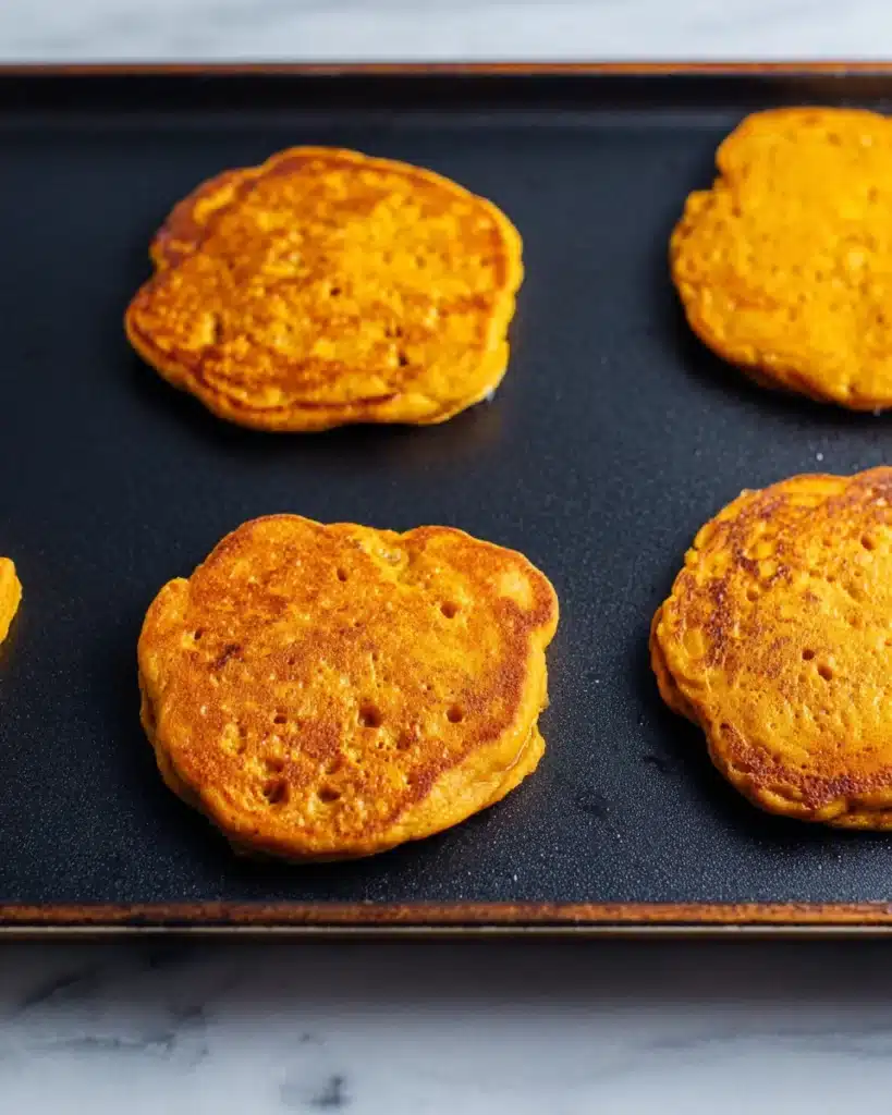 Pumpkin pancakes cooking on a griddle with golden edges