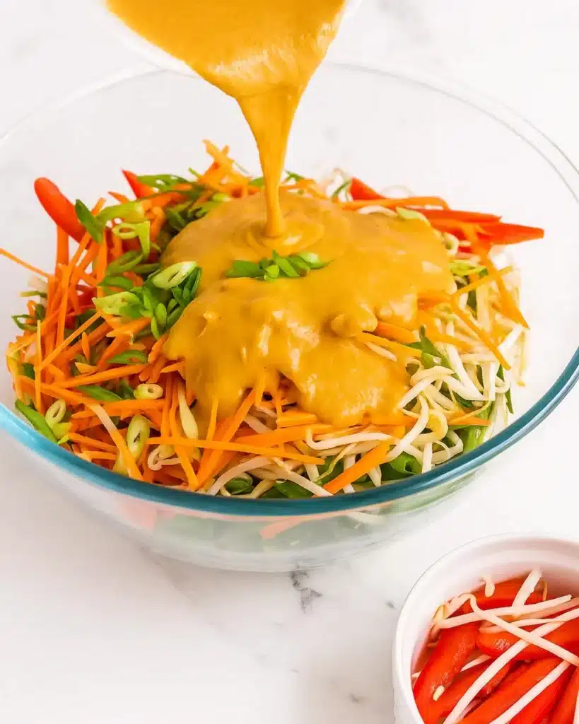 Sesame peanut dressing poured over fresh vegetables and noodles in a bowl.