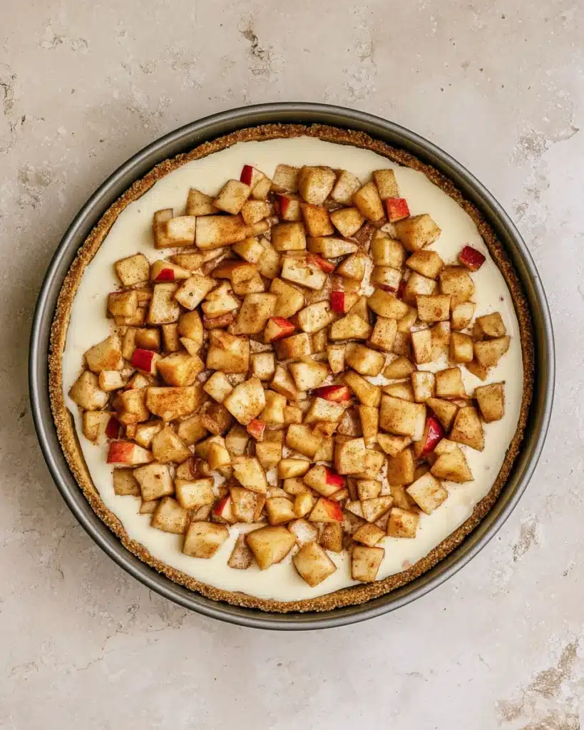 Unbaked apple crumble cheesecake topped with diced spiced apples in pan