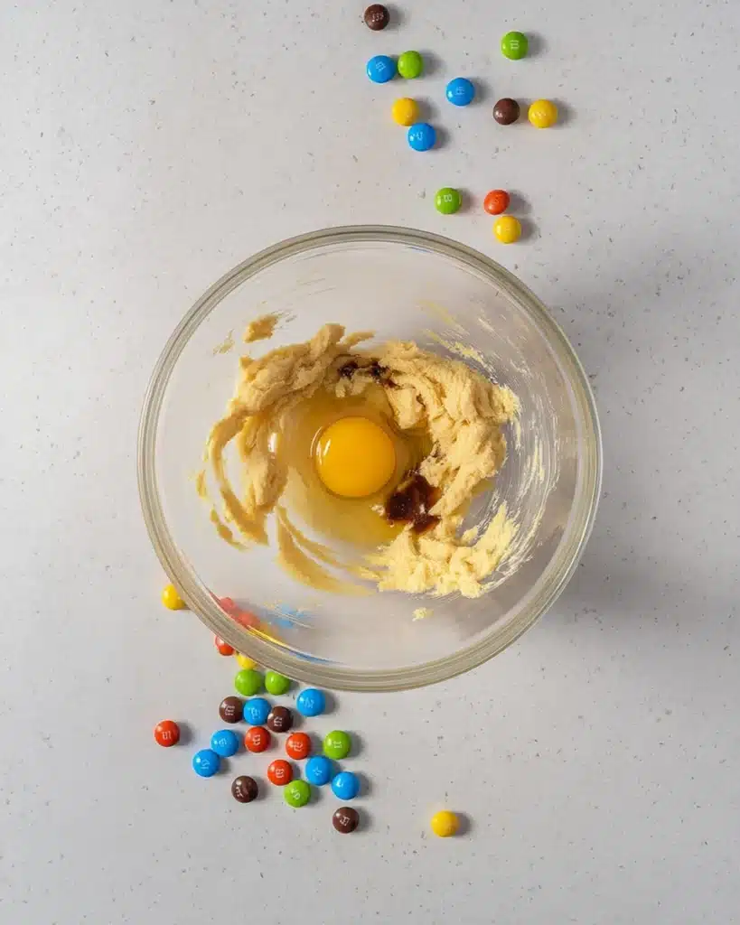 Crumbl Monster Cookie Recipe – Soft & Packed with M&M’s 2 Adding egg and vanilla to Crumbl Monster cookie dough in glass bowl