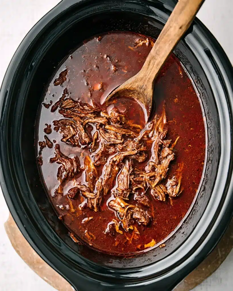 Slow Cooker Birria (Tender & Flavorful Mexican Beef Stew) 3 Shredded birria beef soaking in red broth inside a slow cooker