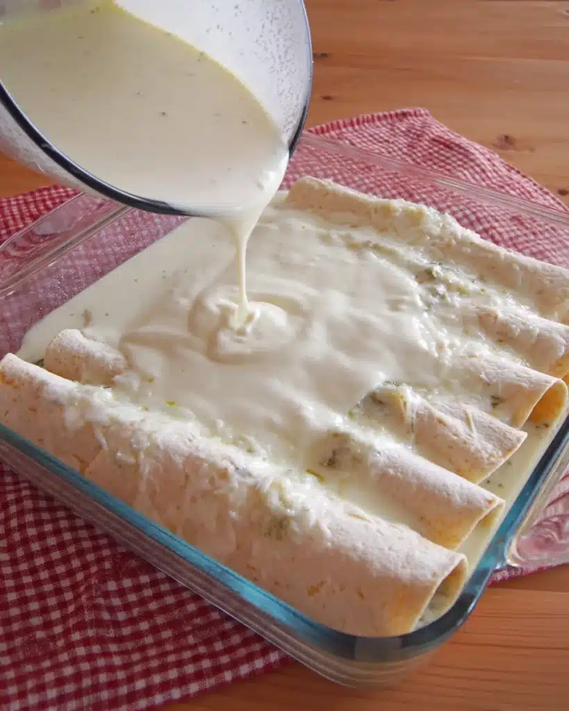 Pouring creamy white sauce over rolled chicken enchiladas in a baking dish