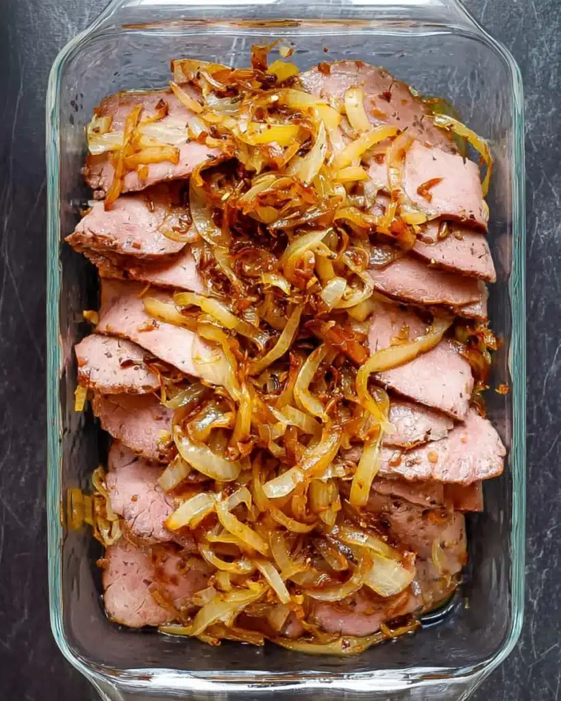 Roast beef layered with caramelized onions in a glass baking dish for French Dip Sliders