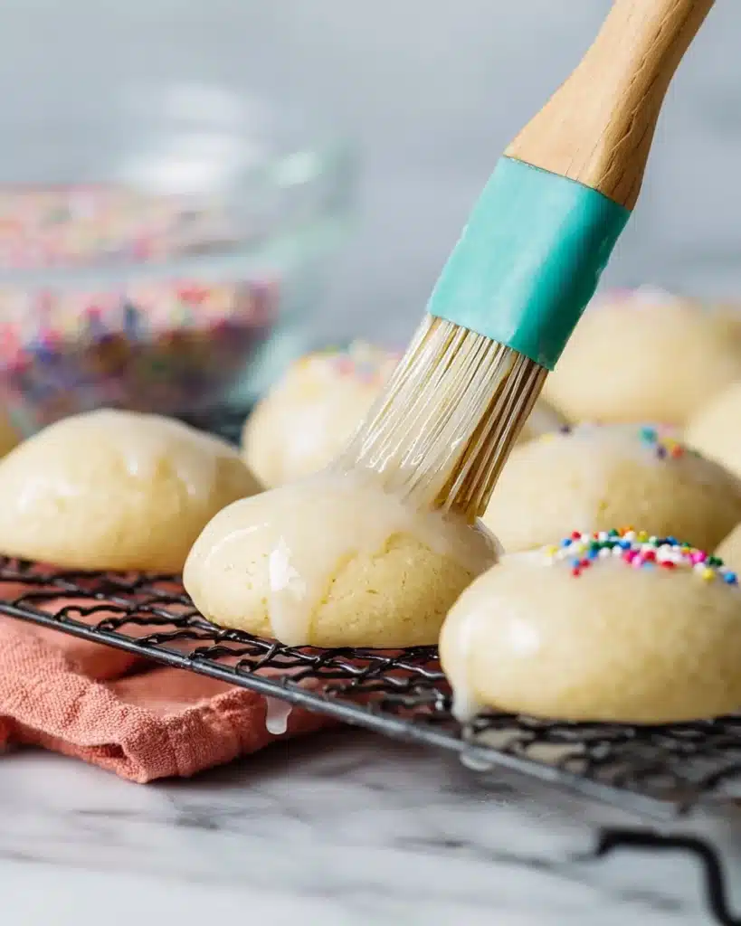 Italian Cookies 3 Pastry brush glazing soft Italian cookies on a cooling rack with sprinkles in the background.