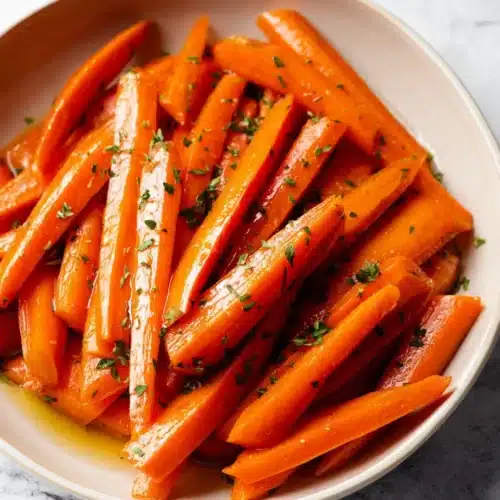 Simple Glazed Carrots 5 Simple Glazed Carrots sticks in a cream bowl topped with fresh herbs on a light stone surface.