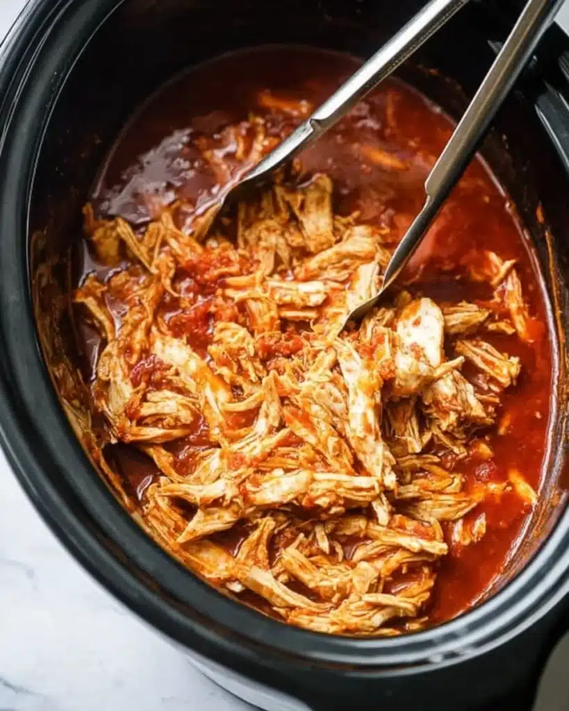 Shredded chicken in slow cooker with red sauce and tongs