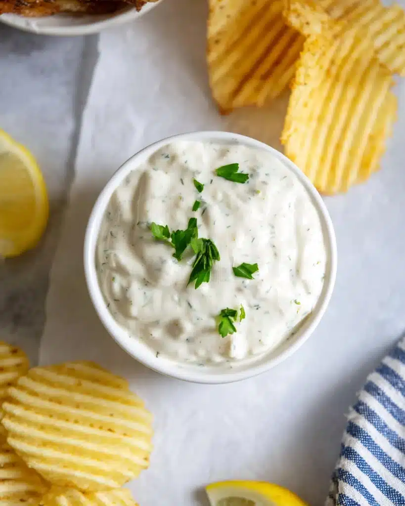 Creamy homemade tartar sauce in a white bowl with lemon and chips