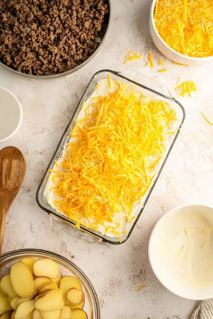 Layered Hobo Casserole topped with shredded cheddar cheese before baking