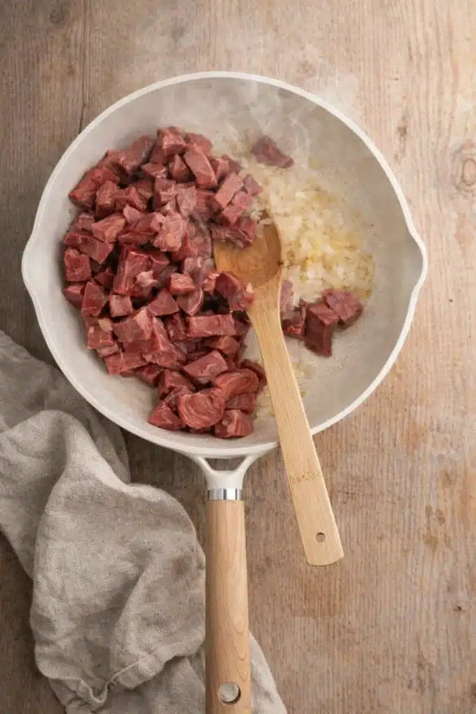 Beef tips and onions cooking in white skillet with wooden spoon