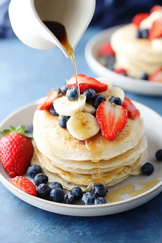 Pouring syrup over fluffy protein pancakes with strawberries, blueberries, and banana
