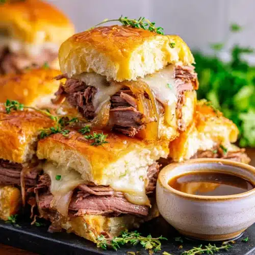 Stacked French dip sliders with roast beef, melted Swiss cheese, and au jus on slate board