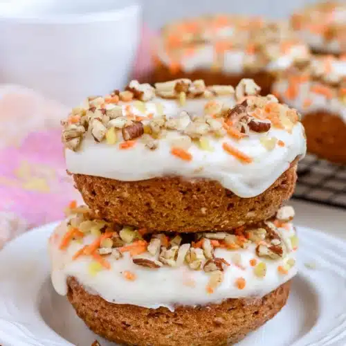Carrot Cake Donuts That Are Soft, Spiced, and Perfect for Easter 4 Stacked carrot cake donuts with cream cheese frosting, chopped nuts, and orange sprinkles on a white plate