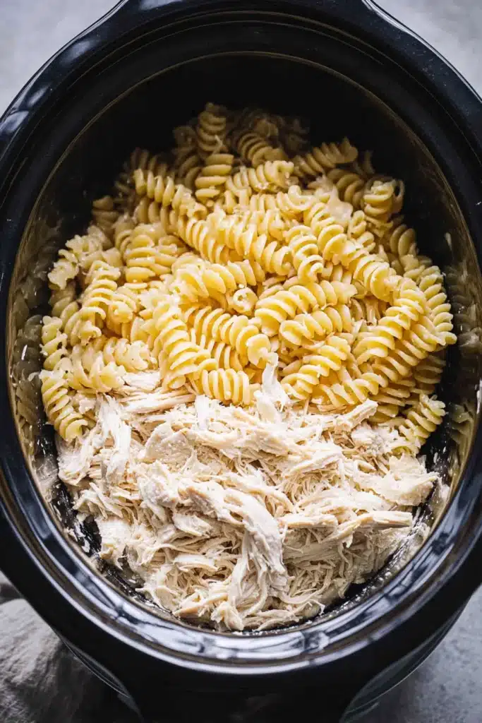 Crockpot garlic parmesan chicken pasta with rotini added to shredded chicken in slow cooker