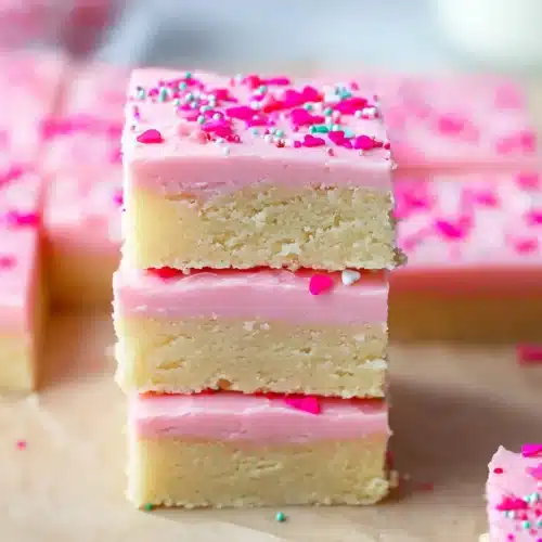 The Best Sugar Cookie Bars 5 Soft sugar cookie bars with pink frosting and sprinkles stacked on parchment paper