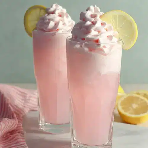 Whipped pink lemonade with creamy topping, ice cubes, and fresh lemon garnish in tall glasses
